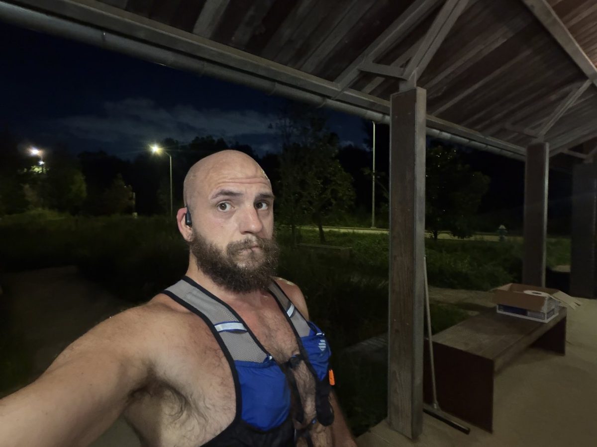 Tank under a park pavilion before sunrise, sweating after the first run back, wearing a blue running vest with a lit path and trees behind him.
